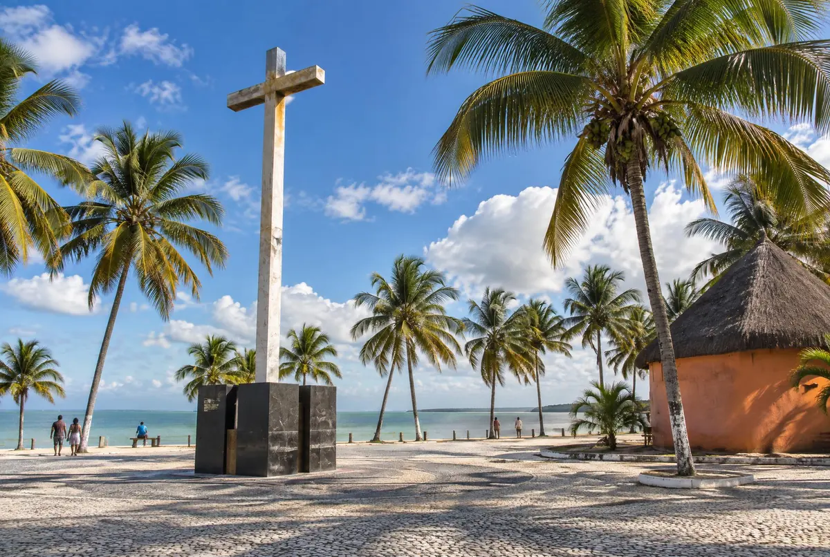 Cruz alta e clara sobre pedestal escuro em praça de pedra à beira-mar, com coqueiros e uma cabana redonda de cobertura de palha.