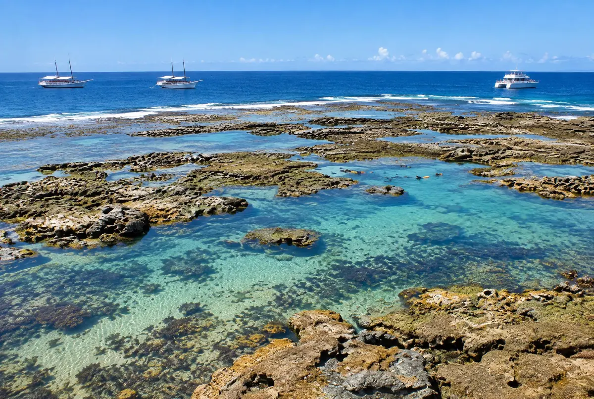 Vista alta da plataforma de recife exposta, com piscinas turquesa e escunas ancoradas ao longe.